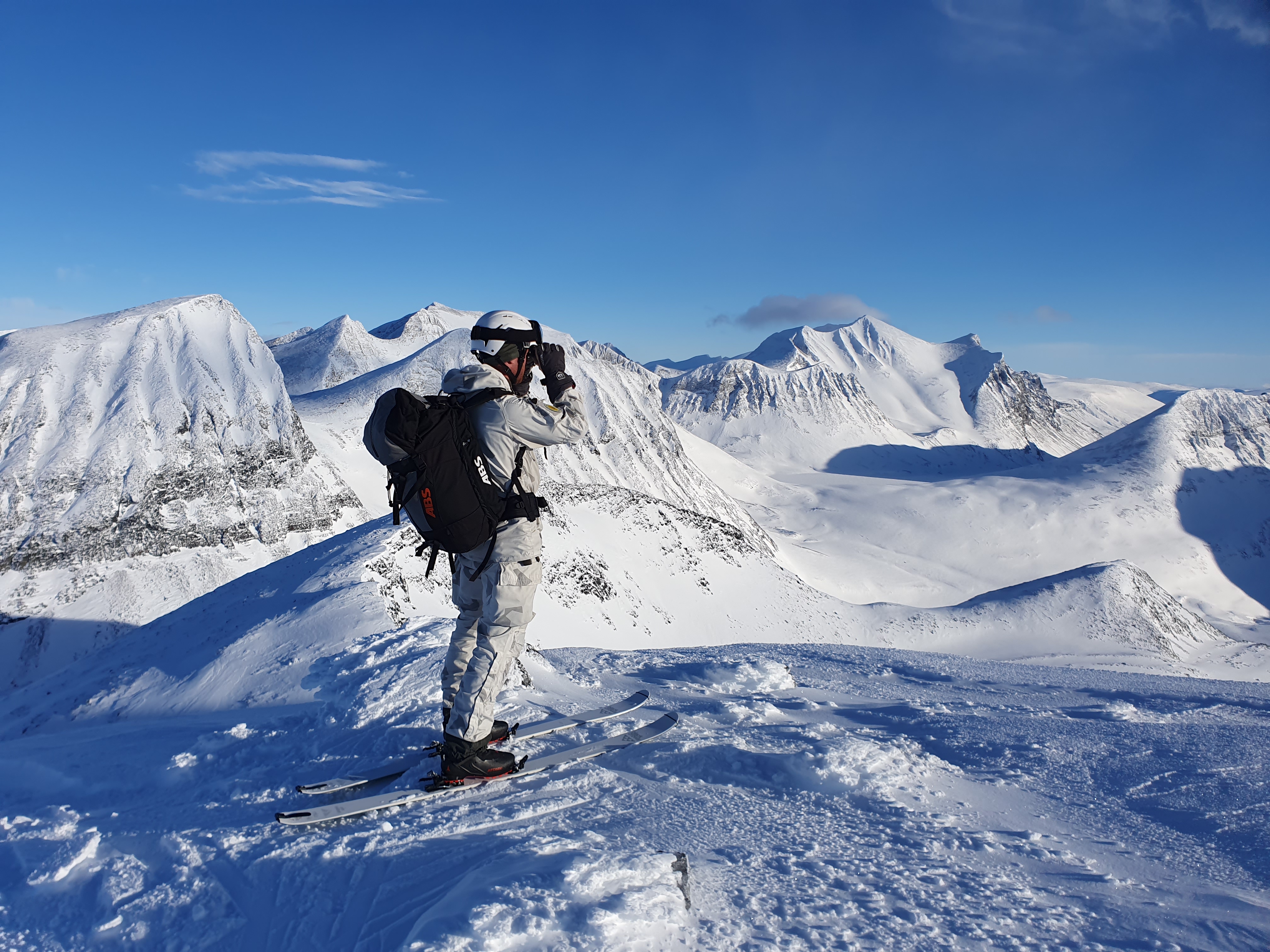 Vitklädd person med ryggsäck och skidor på fötterna står och tittar ut över snöklädda fjäll.