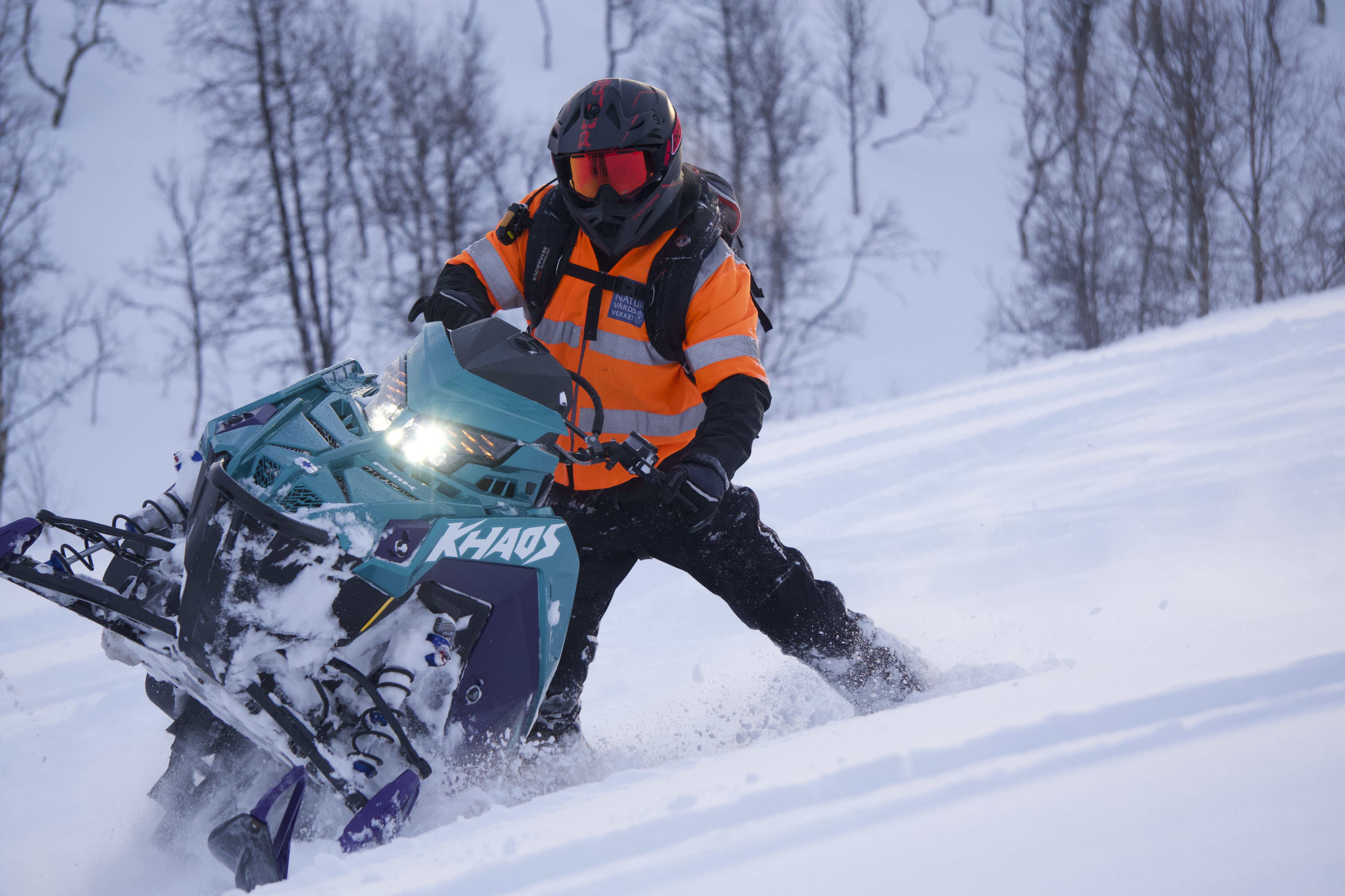 Person med snöskoter lutar den i en snöklädd backe där ena meden är uppe i luften.