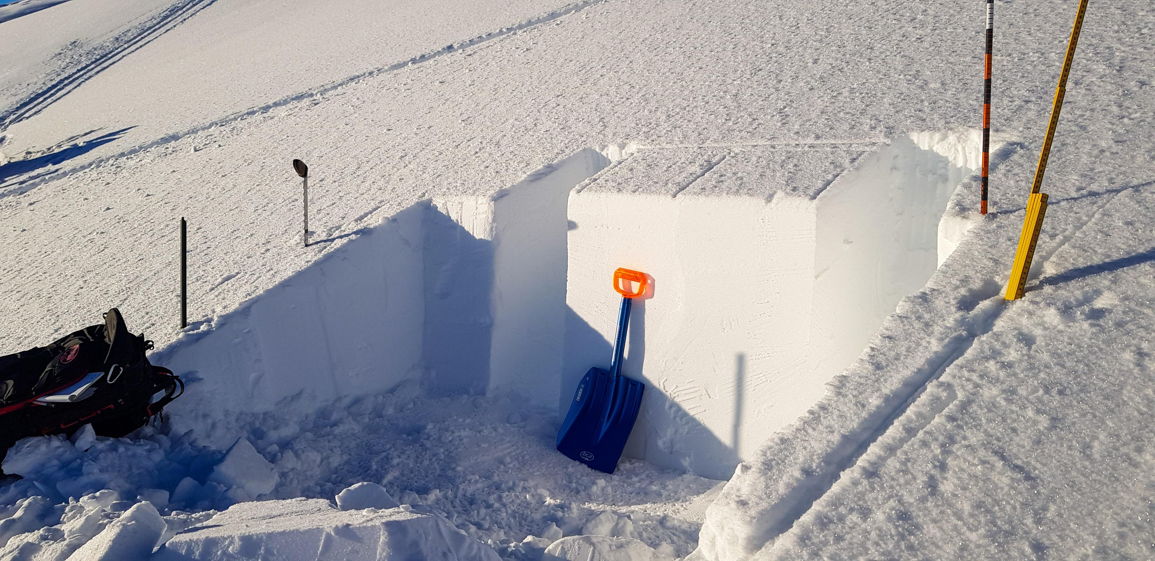 Någon har grävt en grop i snön på ett fjäll. Gropen har raka väggar och man ser en spade och olika mätpinnar bredvid.